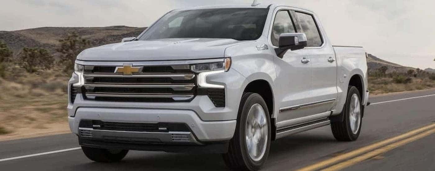 A white 2024 Chevy Silverado 1500 High Country is shown driving on a highway.