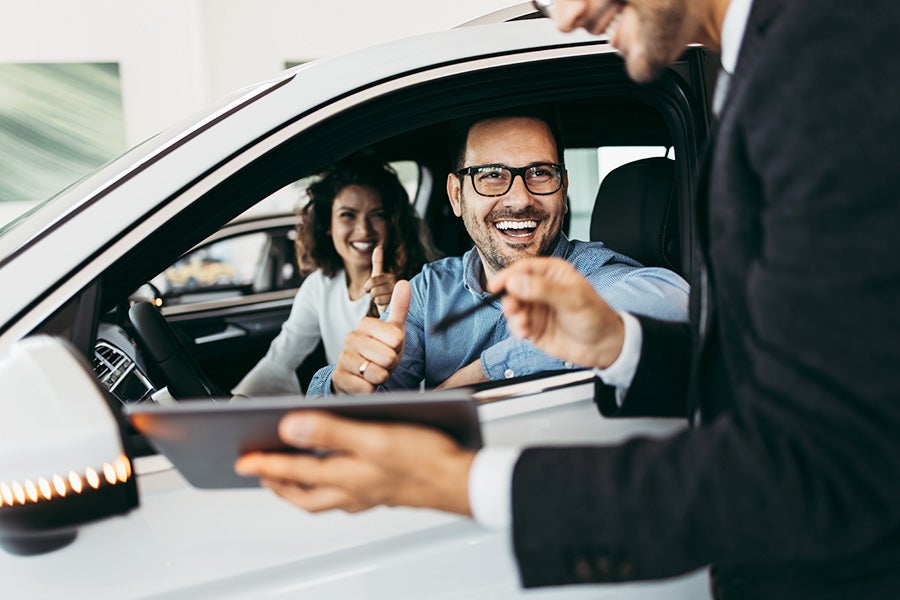 A happy couple in a car gives thumbs up to a salesperson.